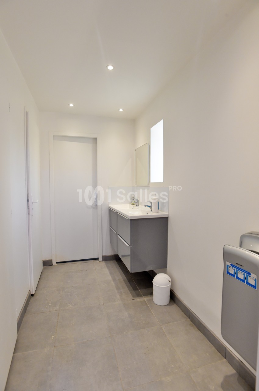 Salle de bain moderne avec carrelage gris, meuble suspendu, miroir, petite fenêtre et poubelle blanche au sol.