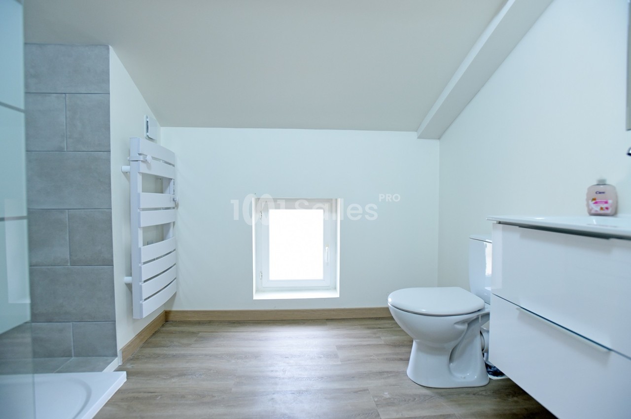 Salle de bain mansardée avec toilettes, meuble blanc, radiateur sèche-serviettes et sol en parquet clair.