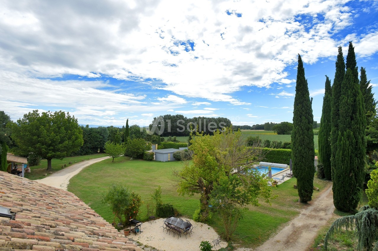 Vue d'un grand jardin avec piscine, allées bordées d'arbres et paysage rural sous un ciel partiellement nuageux.
