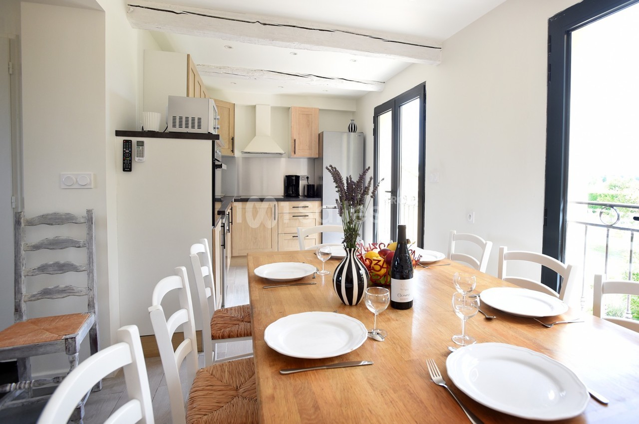 Salle à manger lumineuse avec une grande table en bois dressée, cuisine ouverte en arrière-plan.