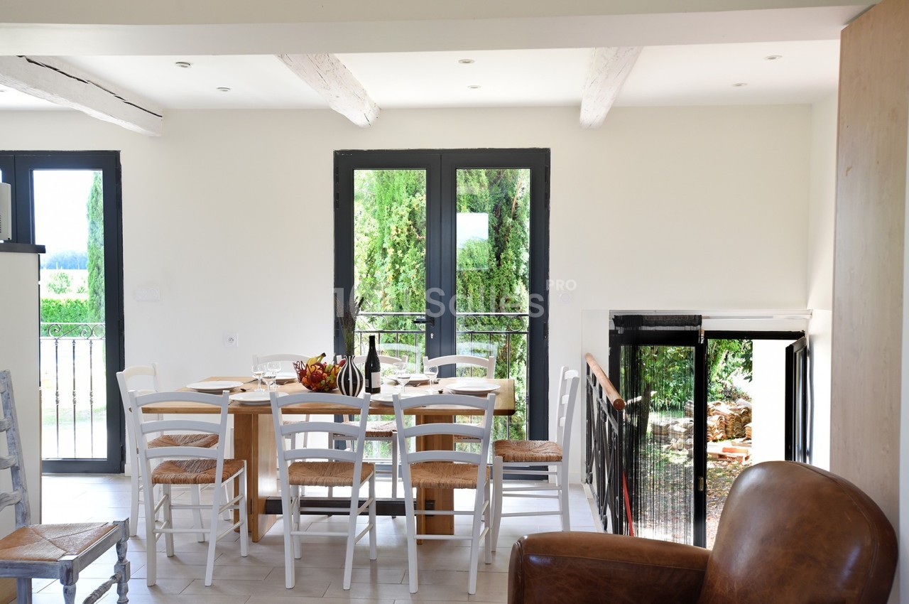 Salle à manger lumineuse avec table en bois, chaises blanches, grandes fenêtres et vue sur un jardin verdoyant.