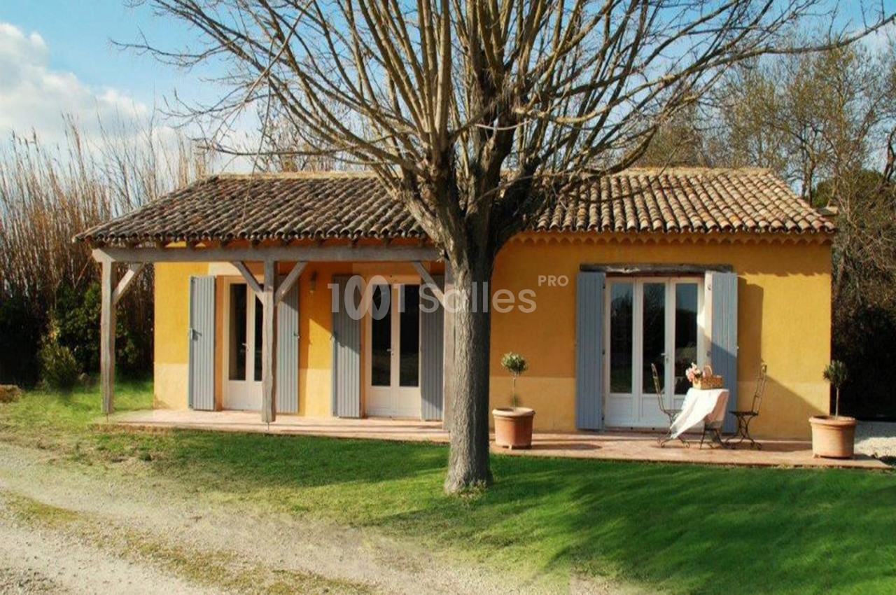 Maison de plain-pied jaune avec volets bleus, terrasse couverte, arbre au premier plan et jardin verdoyant.