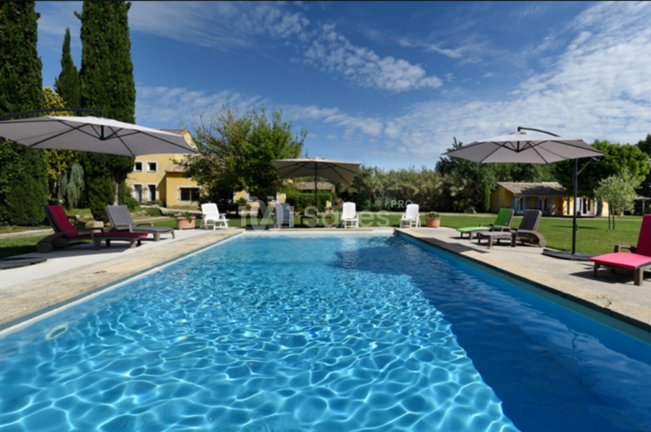 Piscine extérieure entourée de transats et parasols, avec jardin arboré et maison en arrière-plan sous un ciel bleu.