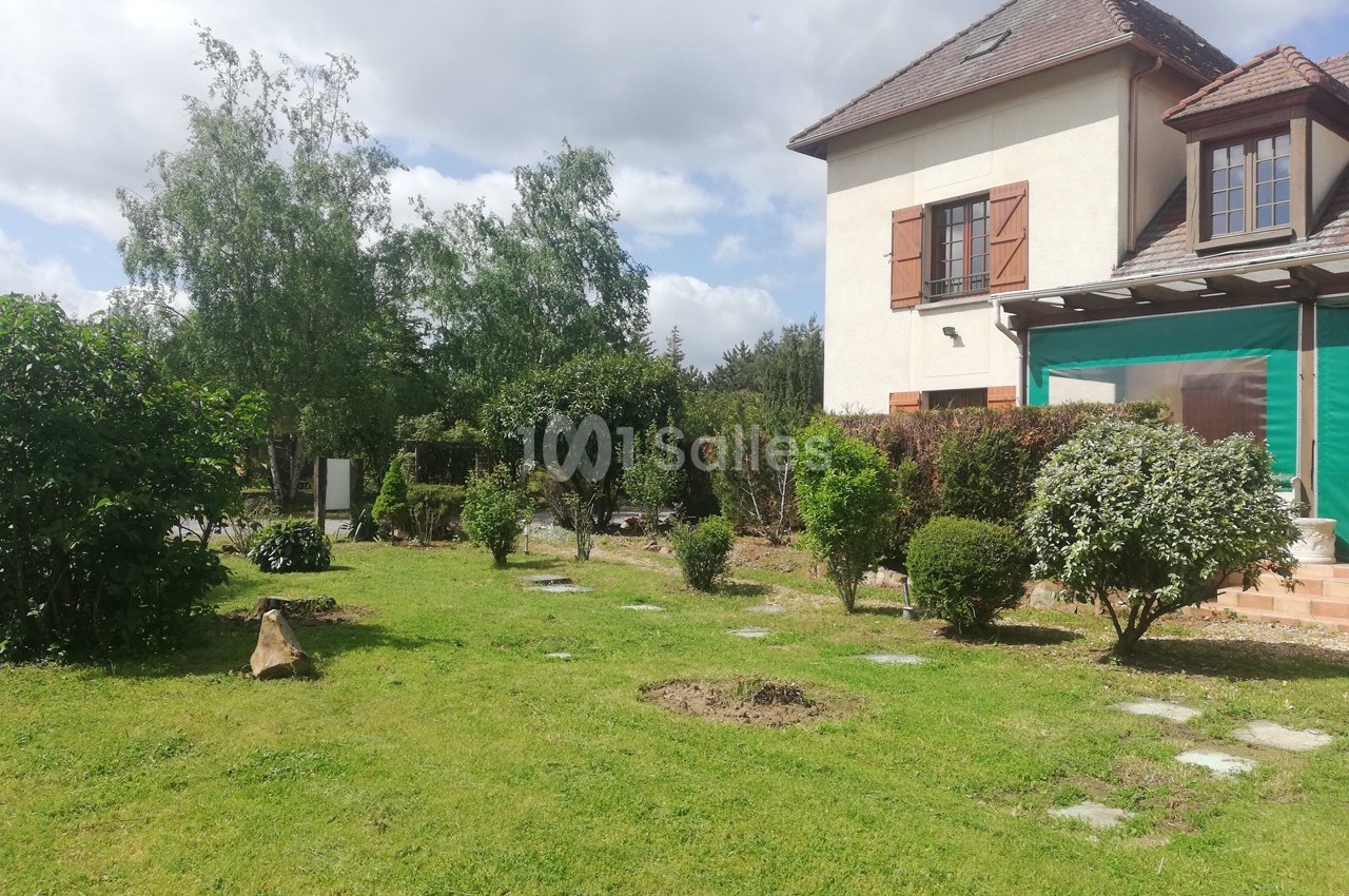 Jardin verdoyant avec pelouse, arbustes et arbres, jouxtant une maison à deux étages avec volets en bois.