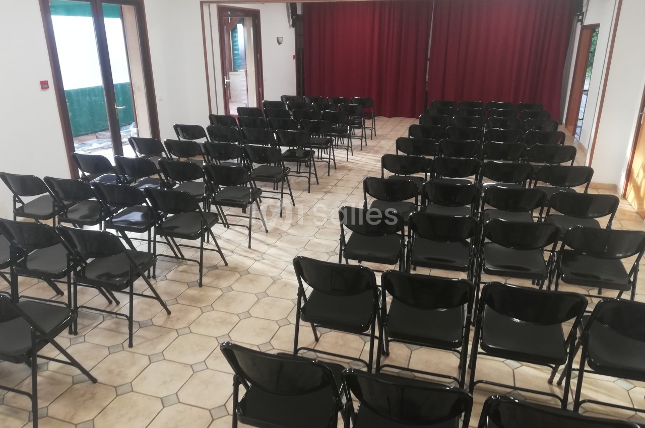Salle avec des rangées de chaises noires pliantes alignées face à une scène avec rideaux rouges.