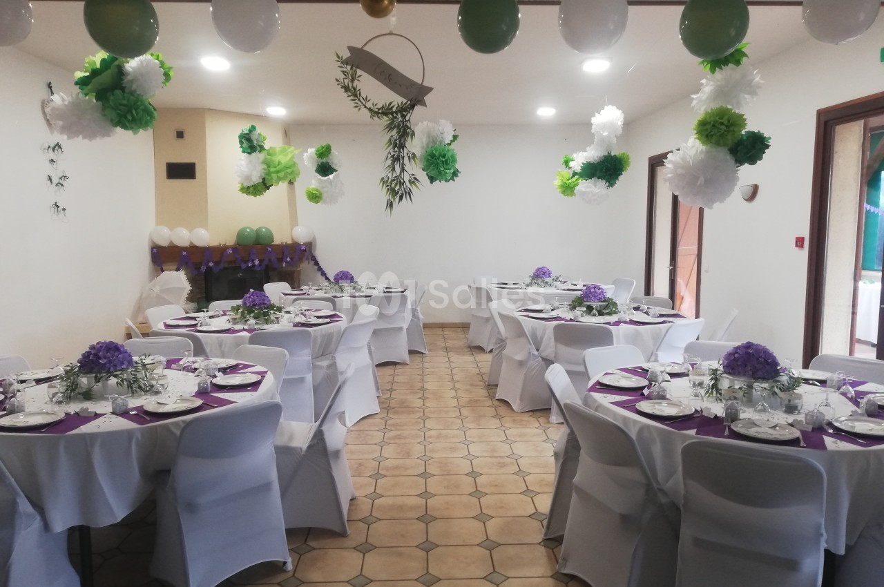 Salle décorée pour un événement avec des tables rondes dressées, des ballons et des fleurs suspendues.