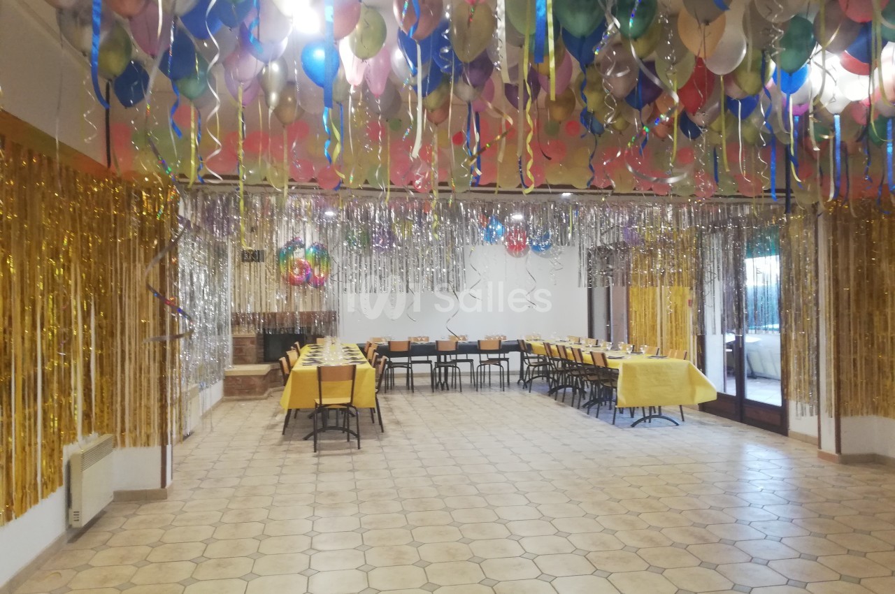 Salle décorée pour une fête avec des ballons colorés au plafond, guirlandes dorées et tables dressées.