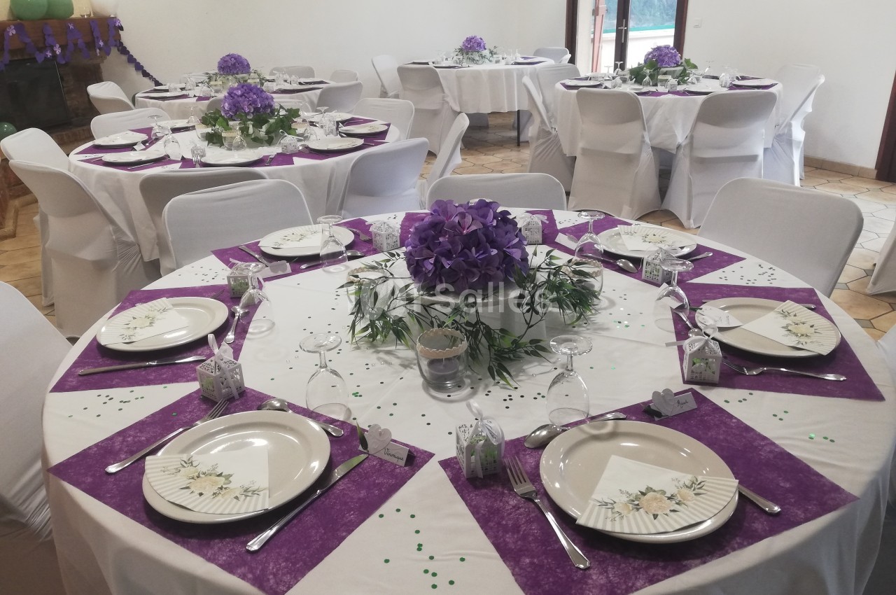 Salle décorée pour un événement avec des tables rondes dressées, nappes blanches et accents violets.
