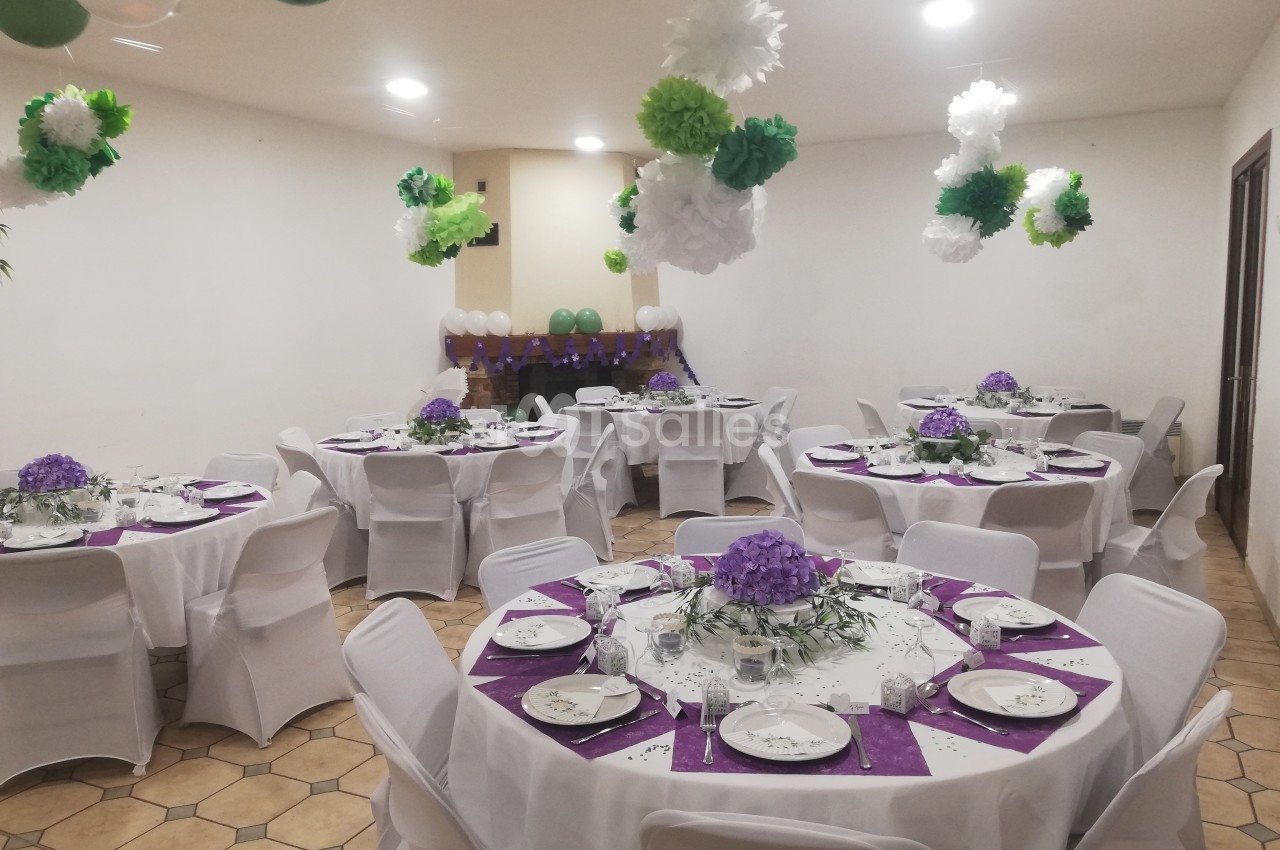 Salle décorée pour un événement avec des tables rondes dressées, nappes blanches, centres de table violets et guirlandes…