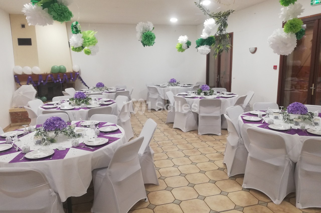Salle décorée pour un événement avec des tables rondes dressées, nappes blanches, centres de table violets et guirlandes…