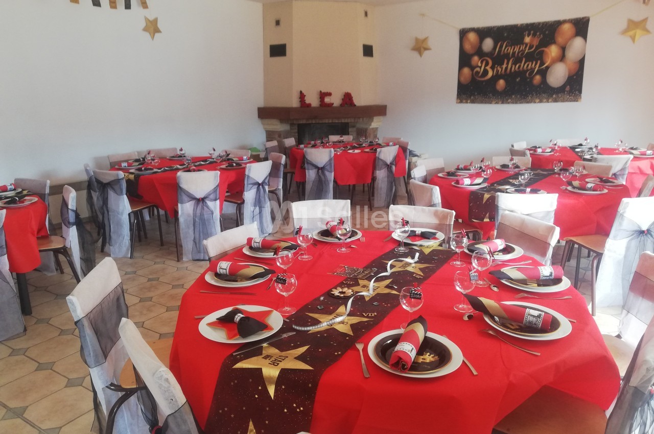 Salle décorée pour un anniversaire avec des tables rondes dressées, nappes rouges et décorations festives.