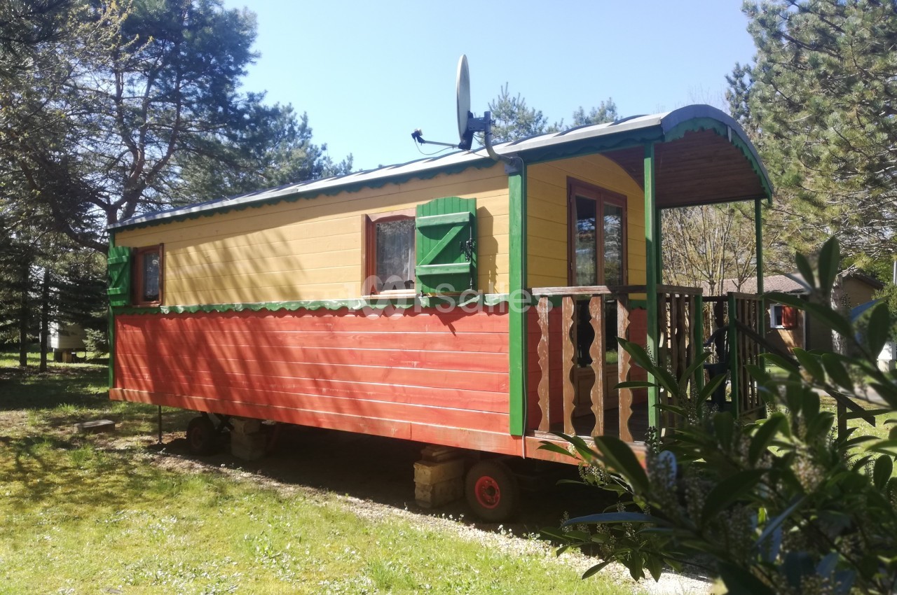 Roulotte en bois rouge et jaune avec volets verts, stationnée sur une pelouse entourée d'arbres.
