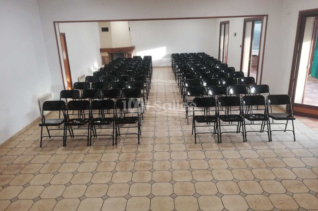 Salle avec des rangées de chaises pliantes noires alignées sur un sol carrelé, éclairée par la lumière naturelle.