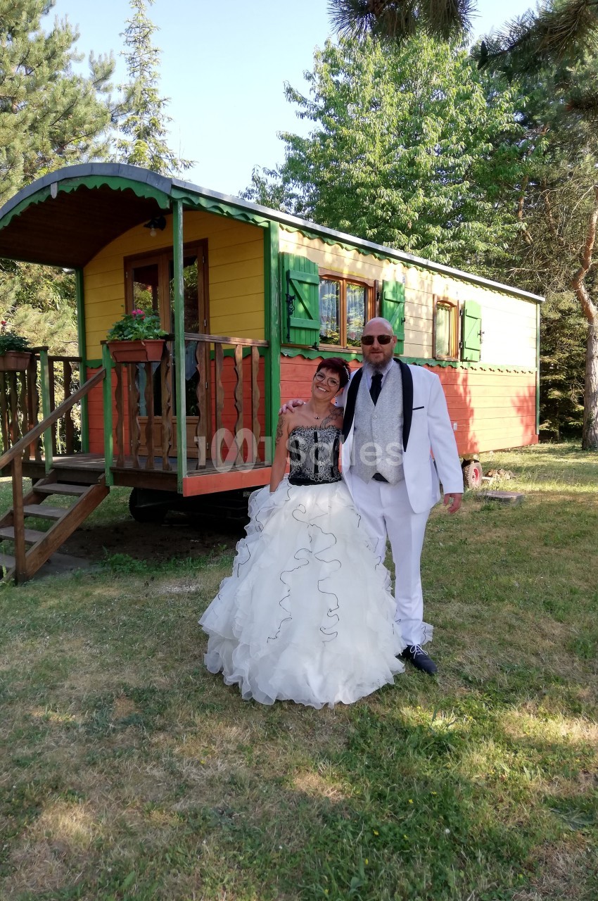 Un couple en tenue de mariage pose devant une roulotte colorée dans un cadre verdoyant.