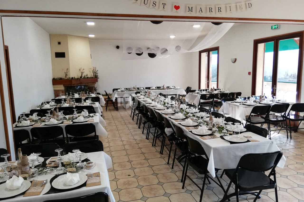 Salle décorée pour un mariage avec des tables alignées, nappes blanches, vaisselle disposée et une bannière ’Just Married’.