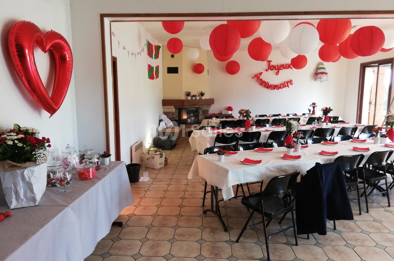 Salle décorée pour un anniversaire avec tables dressées, ballons rouges et blancs, et une guirlande ’Joyeux Anniversaire’.