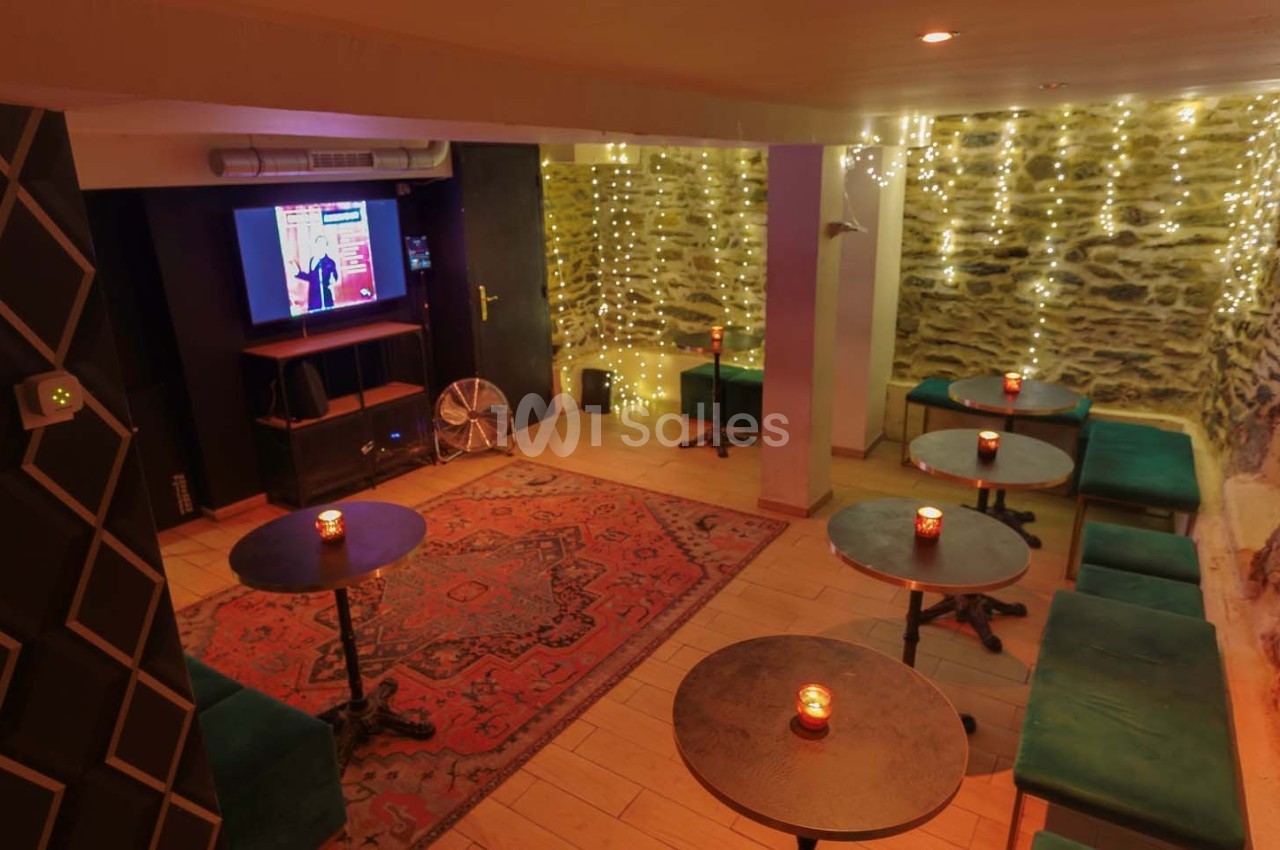 Salle cosy avec tables basses, banquettes, tapis rouge, murs en pierre et guirlandes lumineuses.