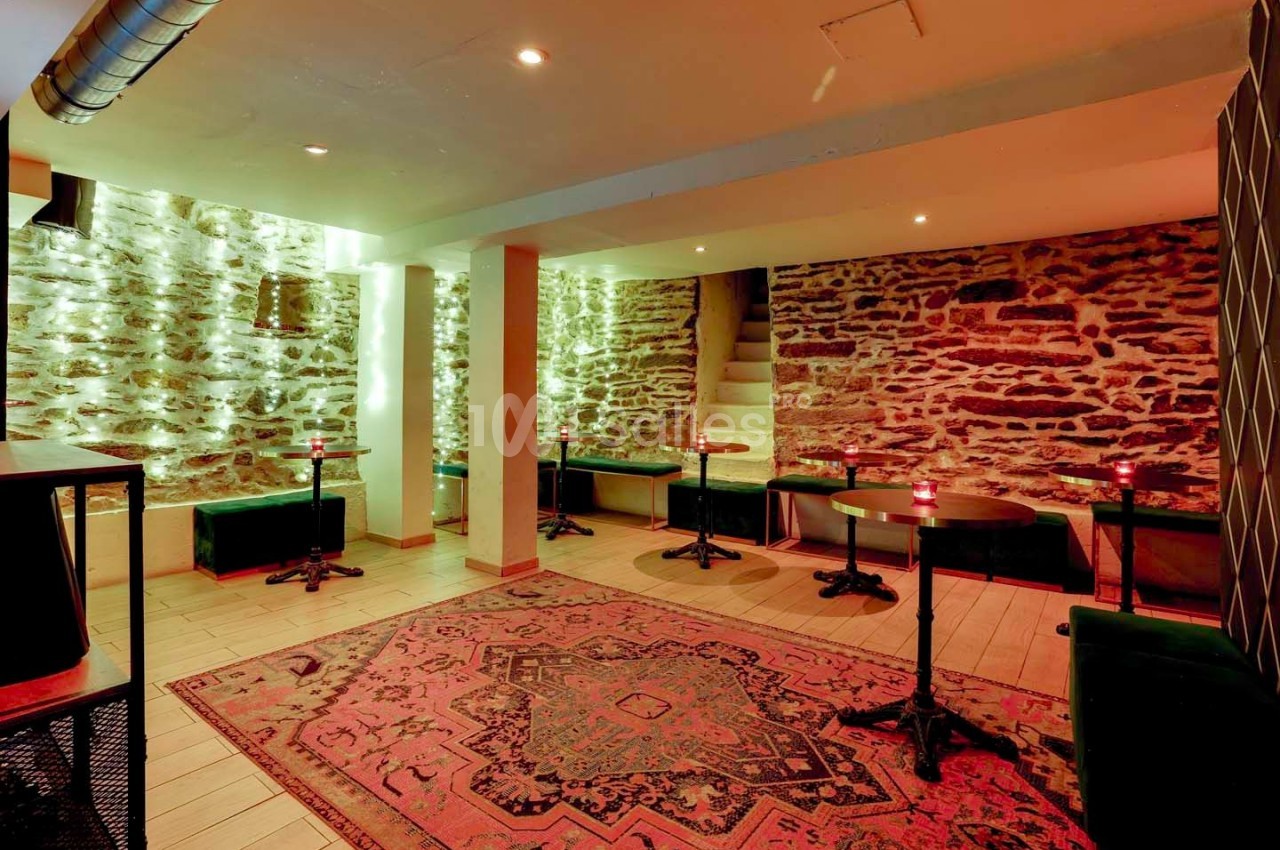 Salle voûtée avec murs en pierre apparente, tables rondes, banquettes vertes et tapis coloré au sol.