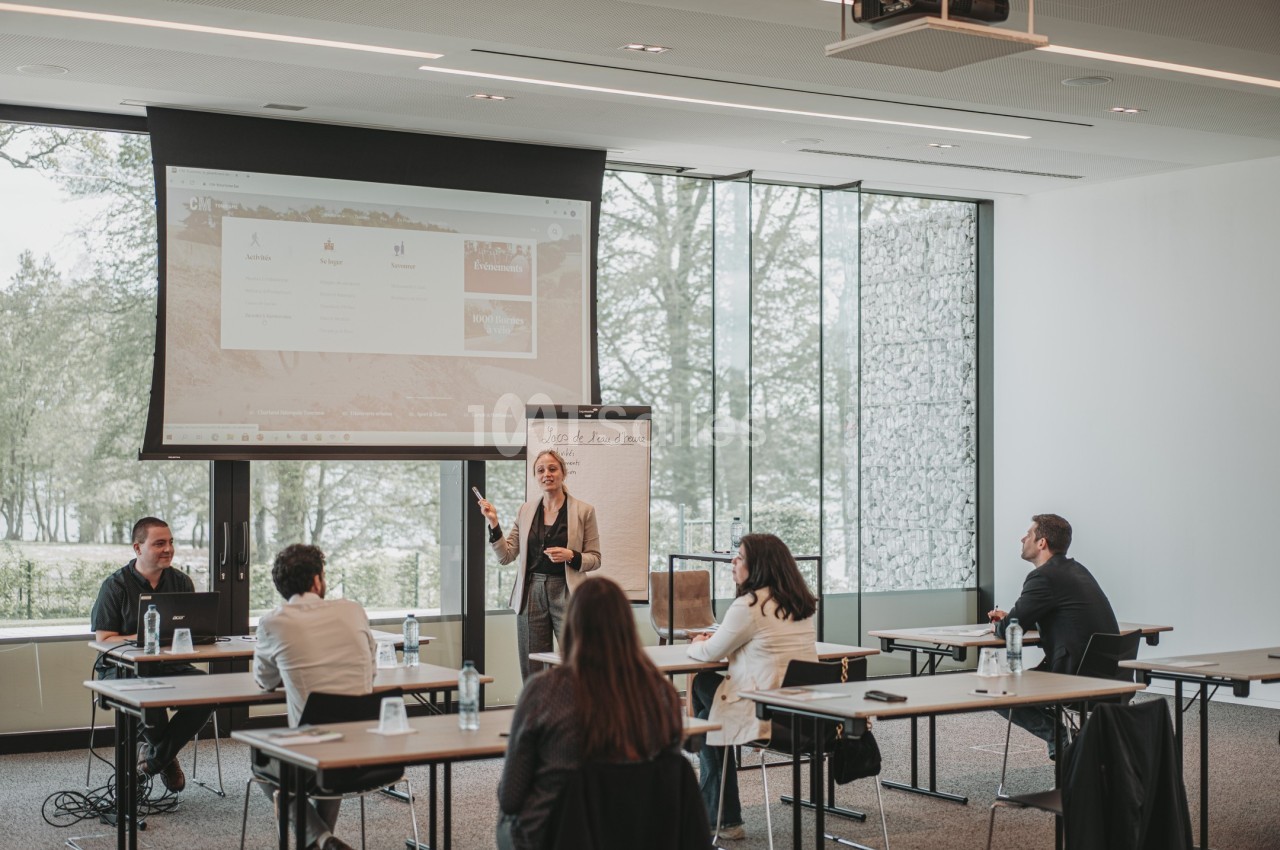 Une femme présente devant un écran et un tableau lors d'une réunion dans une salle lumineuse avec plusieurs participants.