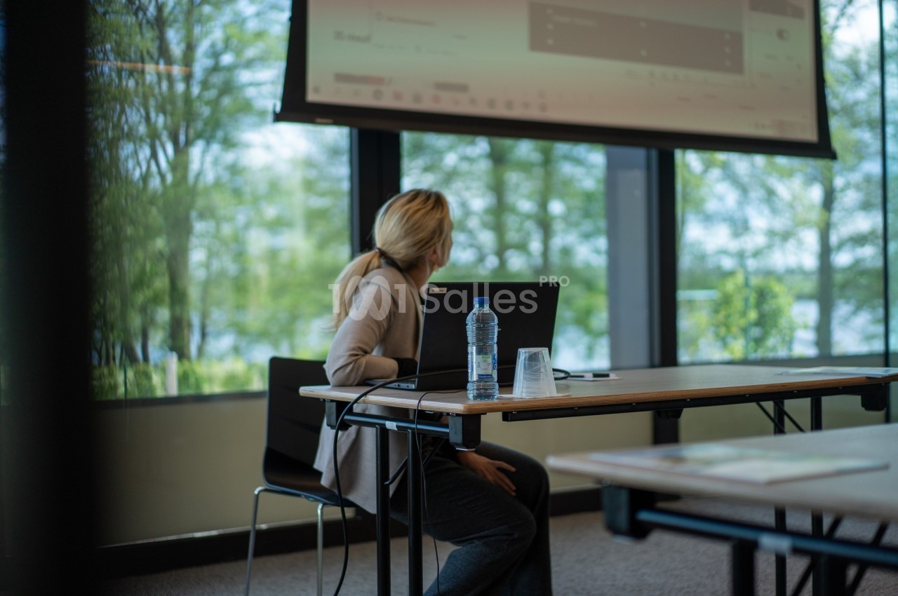 Une femme assise à une table regarde un écran de projection dans une salle lumineuse avec vue sur l'extérieur.