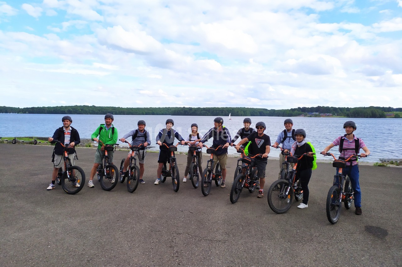 Un groupe de personnes en tenue décontractée posant avec des vélos électriques devant un lac sous un ciel partiellement…