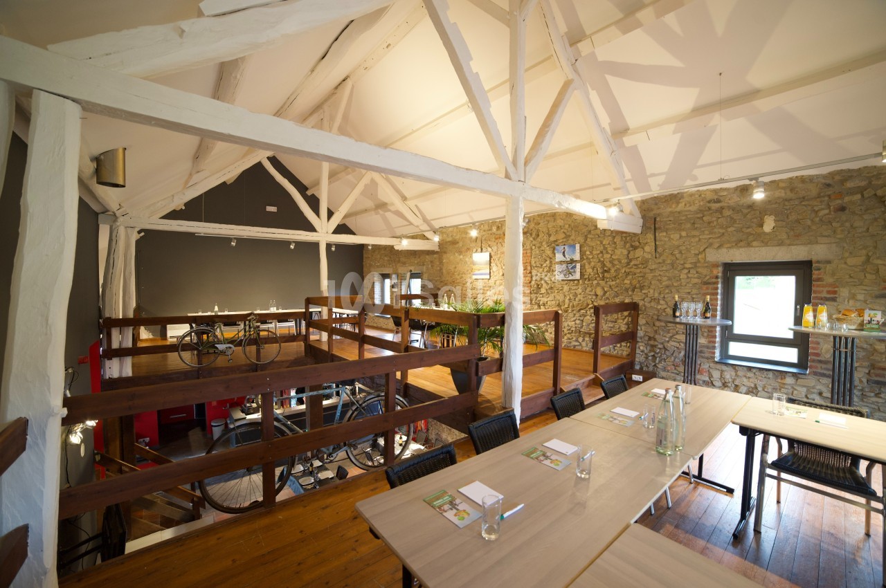 Salle lumineuse avec poutres apparentes, tables en bois, murs en pierre et mezzanine aménagée avec des vélos exposés.
