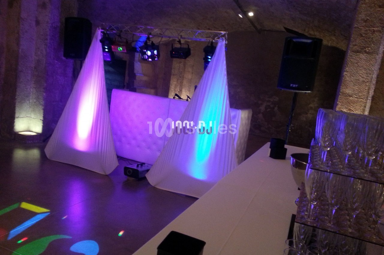 Installation de DJ avec éclairage coloré dans une salle en pierre, table de mixage et verres empilés au premier plan.