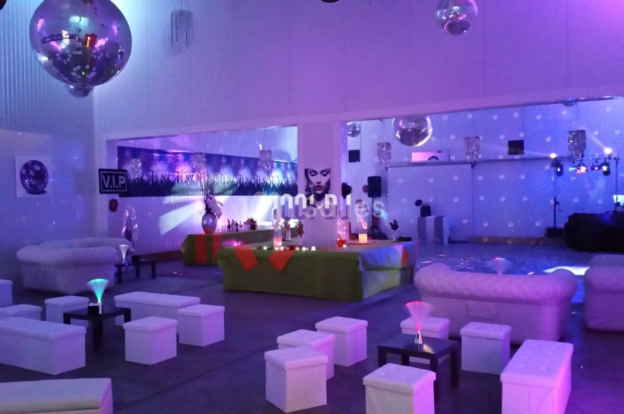 Salle décorée pour un événement, avec canapés blancs, tables basses, éclairage coloré et ambiance festive.