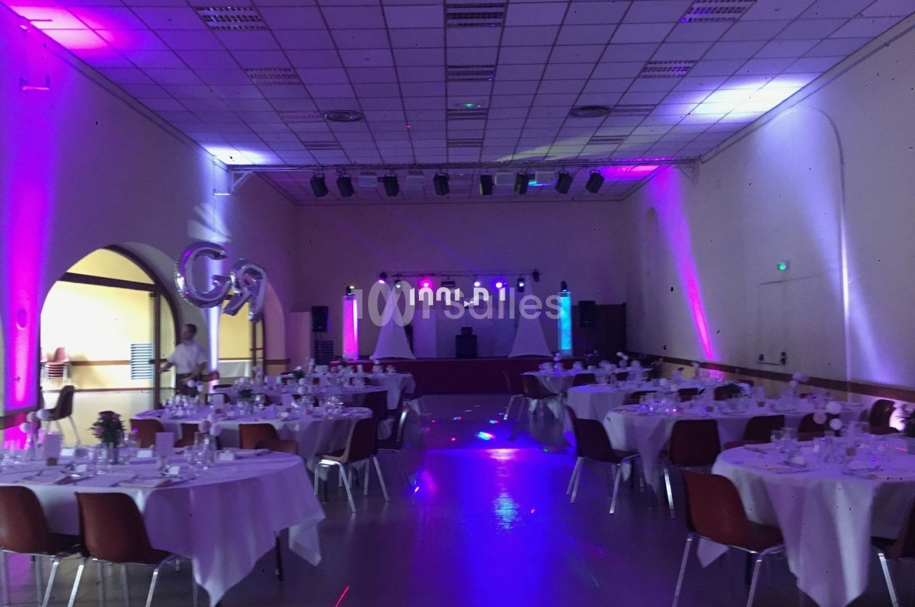 Salle de réception décorée avec des tables rondes dressées et une scène éclairée de lumières colorées.