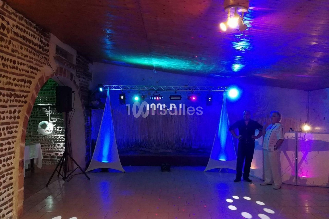 Salle de réception avec éclairage coloré, piste de danse vide et deux personnes debout près d'un bar lumineux.
