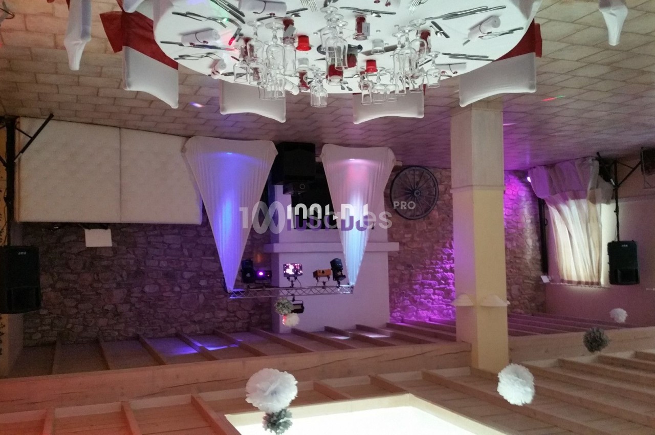 Salle de réception décorée avec des tables dressées, éclairages colorés et espace DJ en arrière-plan.
