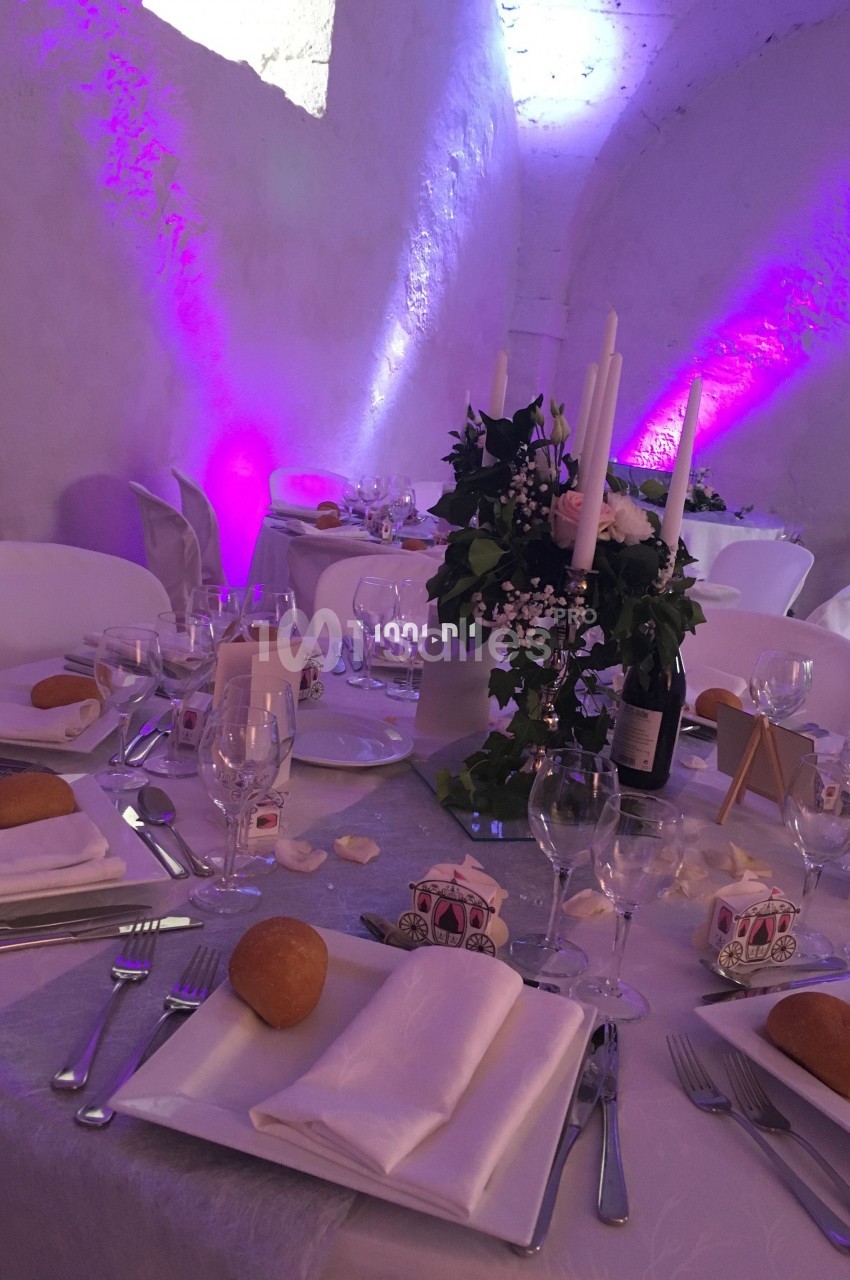 Table de réception décorée avec des bougies, des fleurs et des accessoires de mariage, dans une salle aux lumières violettes.