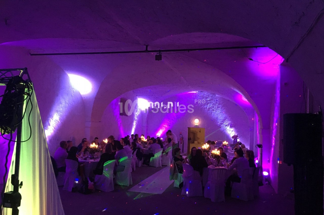 Salle voûtée éclairée de lumières colorées, avec des invités assis à des tables décorées pour un événement festif.