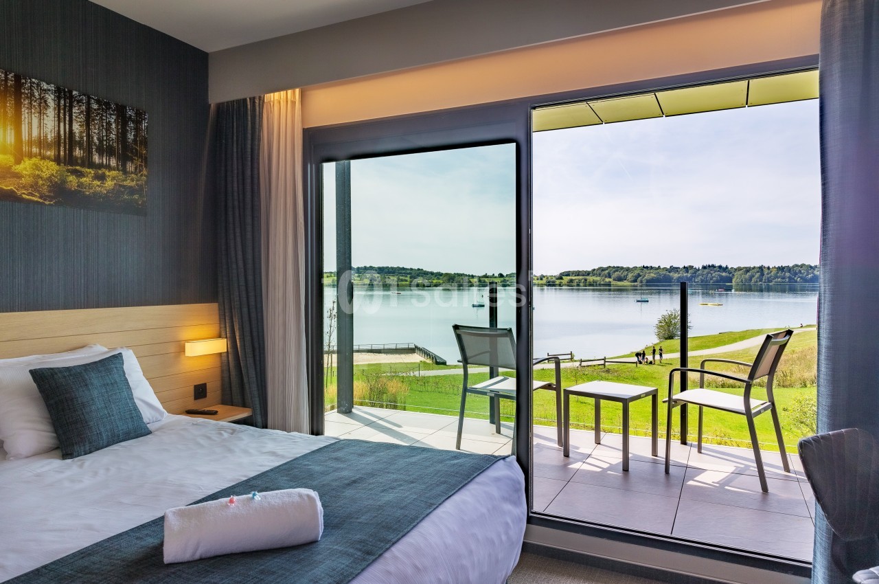 Chambre d'hôtel avec lit double, balcon aménagé et vue sur un lac entouré de verdure.