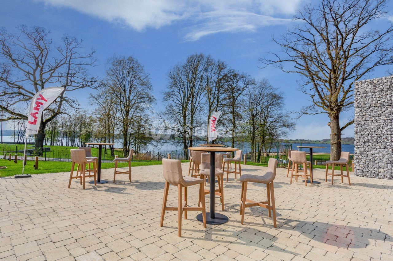 Terrasse extérieure avec tables hautes et chaises, entourée d'arbres et offrant une vue sur un plan d'eau.