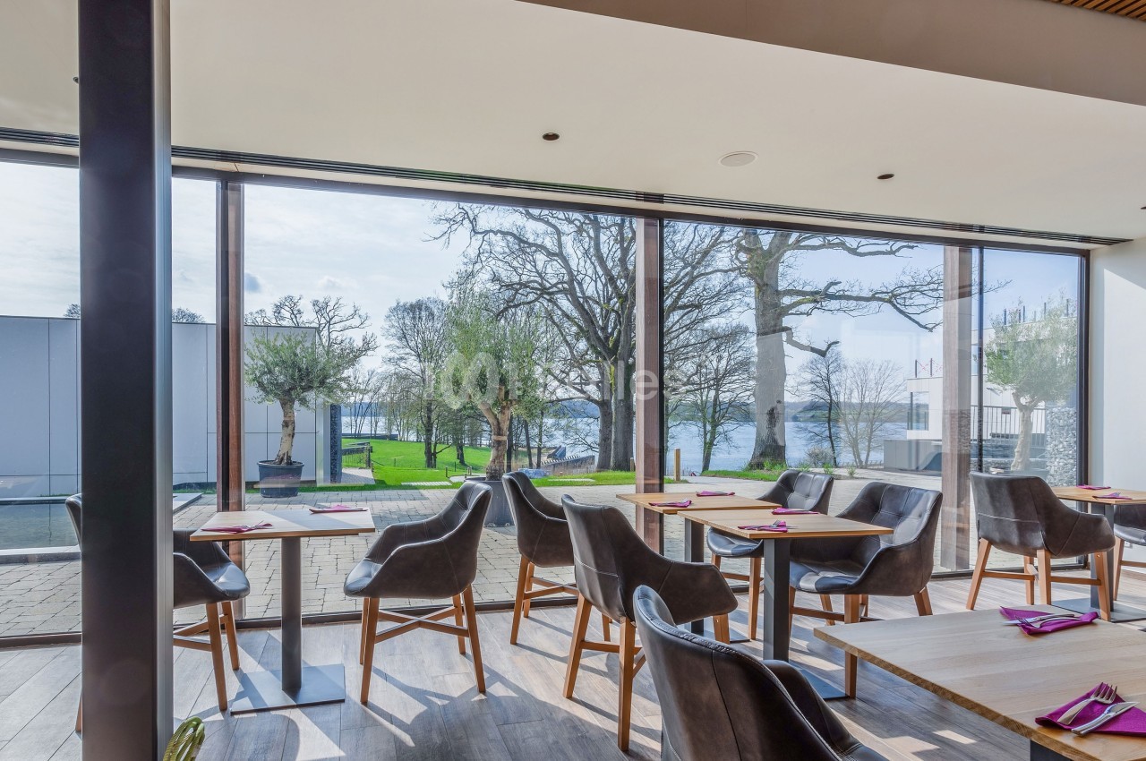 Salle de restaurant lumineuse avec grandes baies vitrées donnant sur un jardin et une vue sur un plan d'eau.