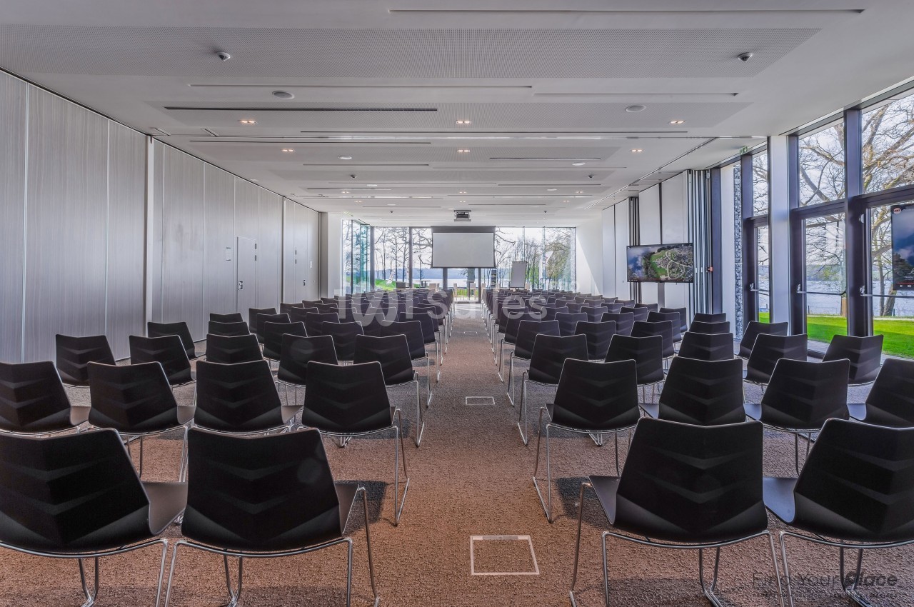 Salle de conférence lumineuse avec rangées de chaises noires, écran de projection et grandes baies vitrées donnant sur un…