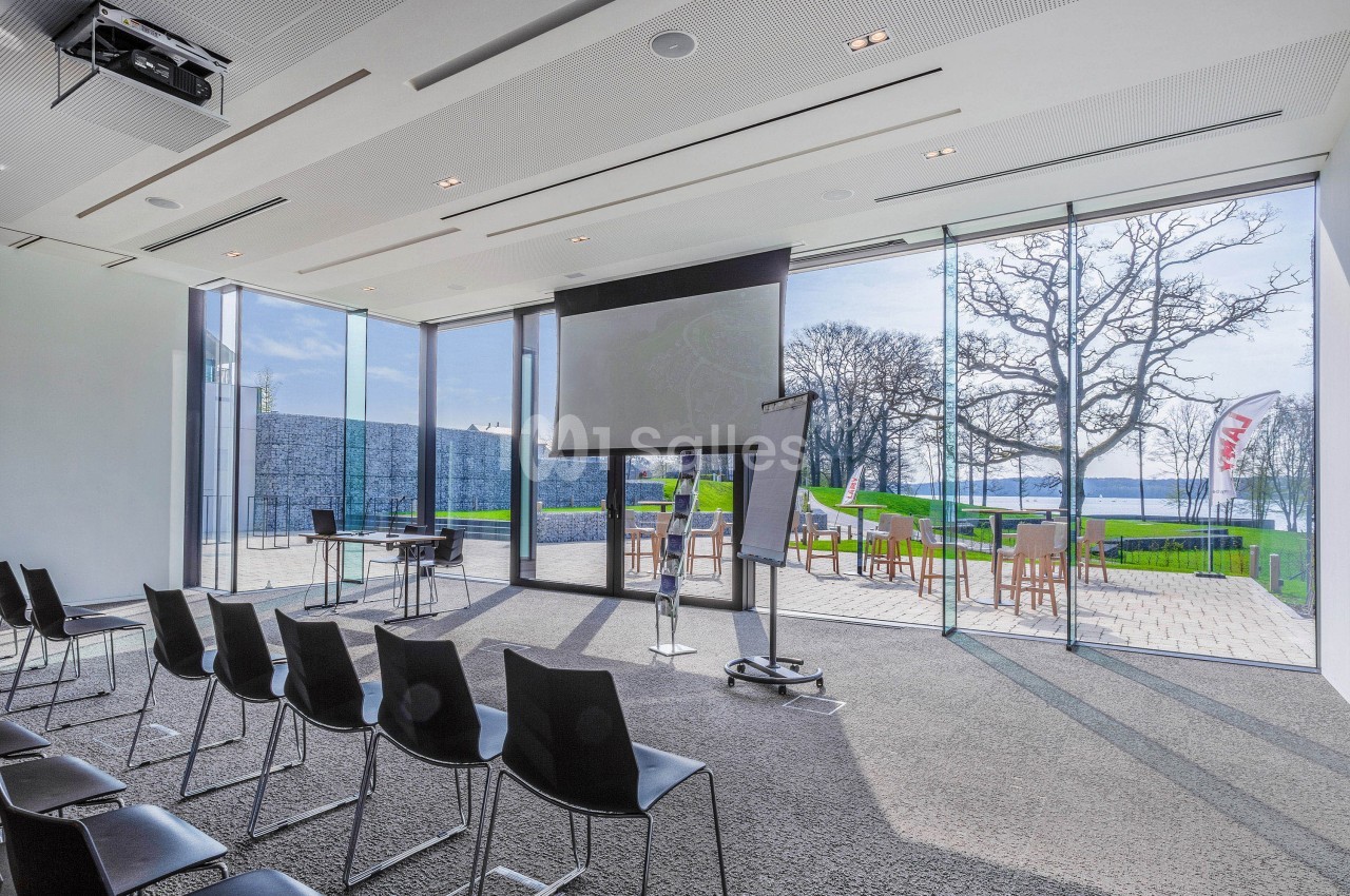 Salle de conférence lumineuse avec chaises alignées, écran de projection et vue sur un espace extérieur verdoyant.