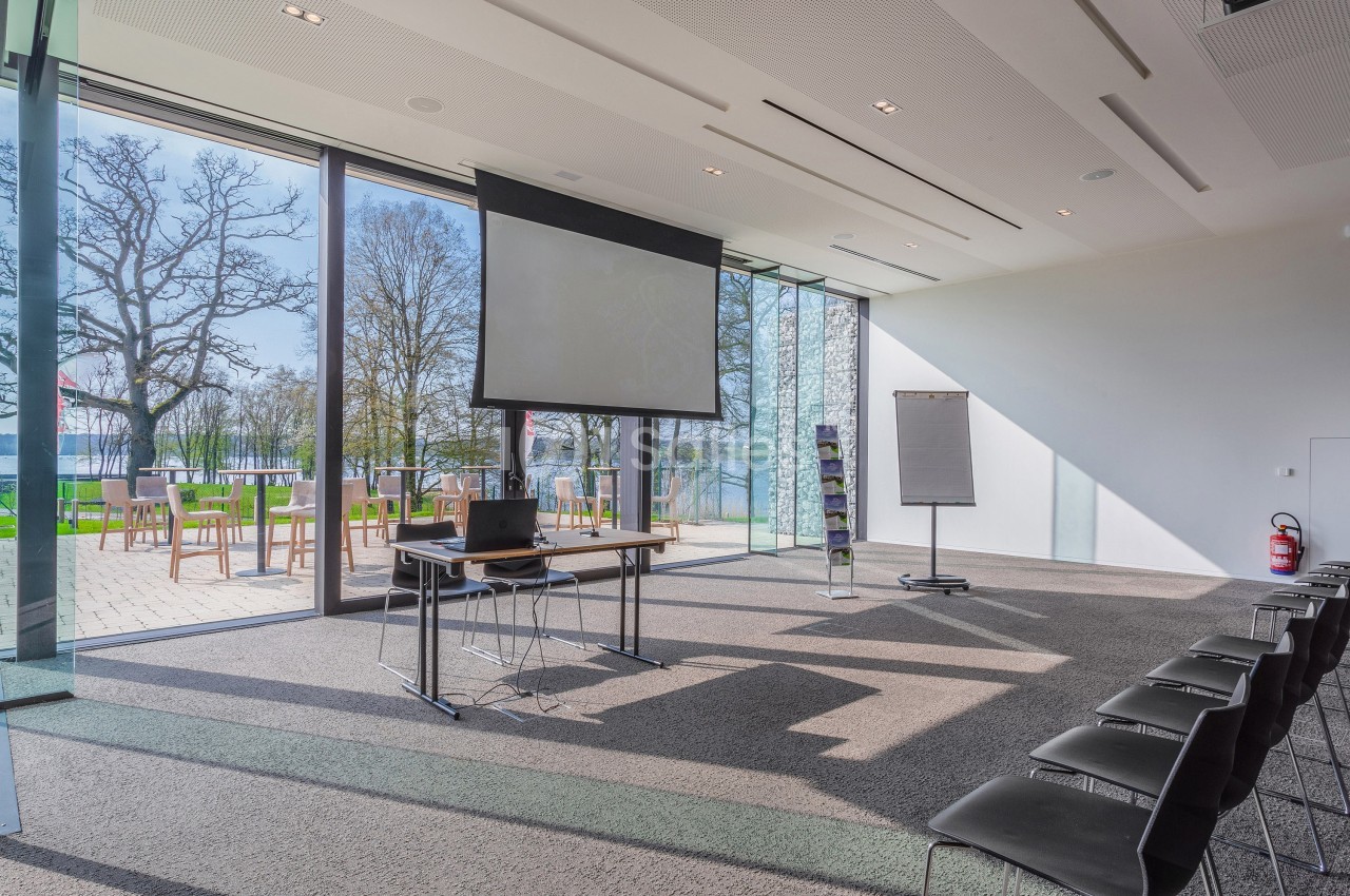 Salle de réunion lumineuse avec chaises alignées, écran de projection, paperboard et vue sur un espace extérieur arboré.
