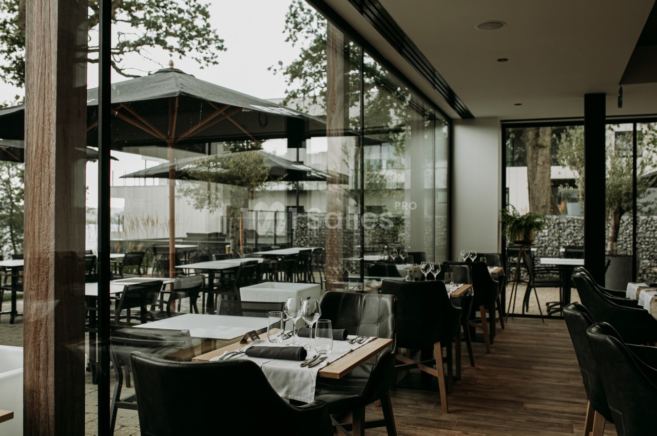Salle de restaurant moderne avec tables dressées, grandes baies vitrées et vue sur une terrasse extérieure arborée.