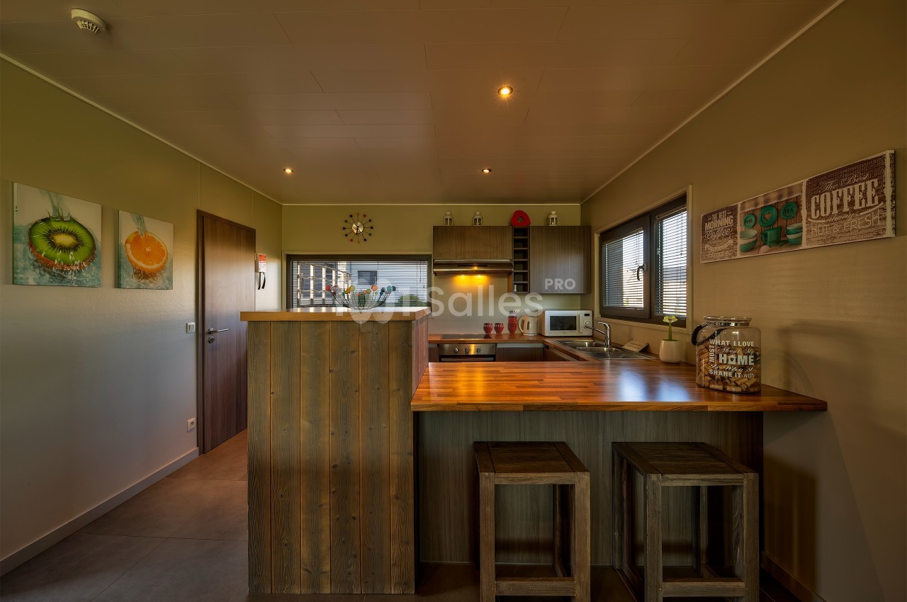 Cuisine moderne avec comptoir en bois, tabourets, électroménagers, décorations murales et éclairage chaleureux.