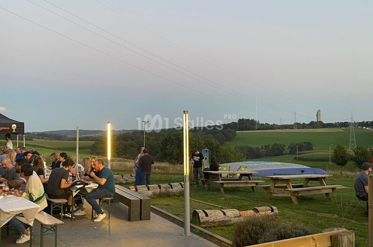 Des personnes dînent en plein air sur des tables en bois, avec une vue sur des collines et un ciel dégagé en arrière-plan.