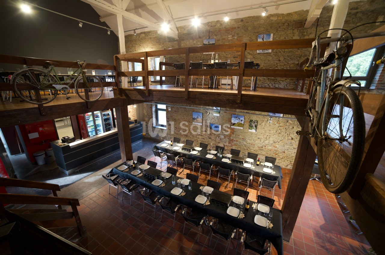 Salle à manger spacieuse avec tables dressées, murs en pierre, mezzanine en bois et vélos accrochés en décoration.