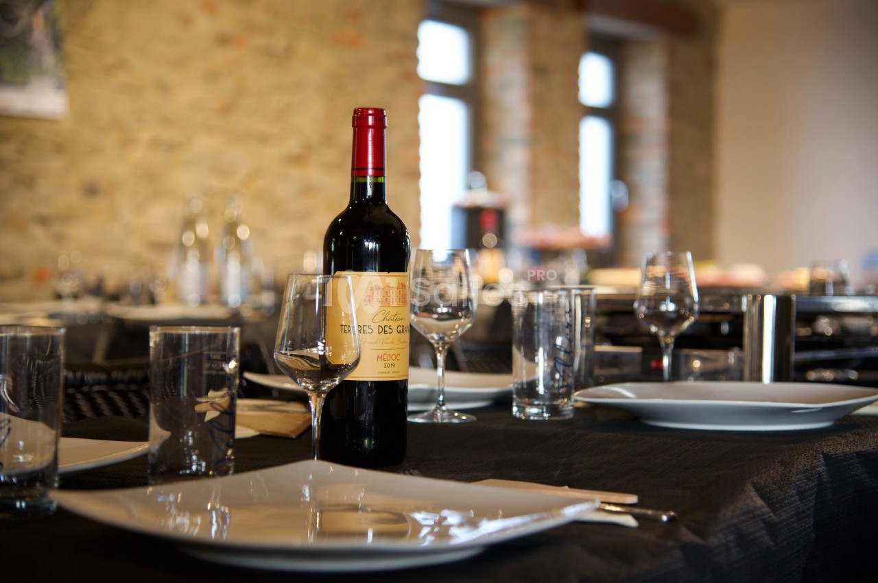 Bouteille de vin rouge posée sur une table dressée avec assiettes, verres et couverts, dans une salle lumineuse en pierre.