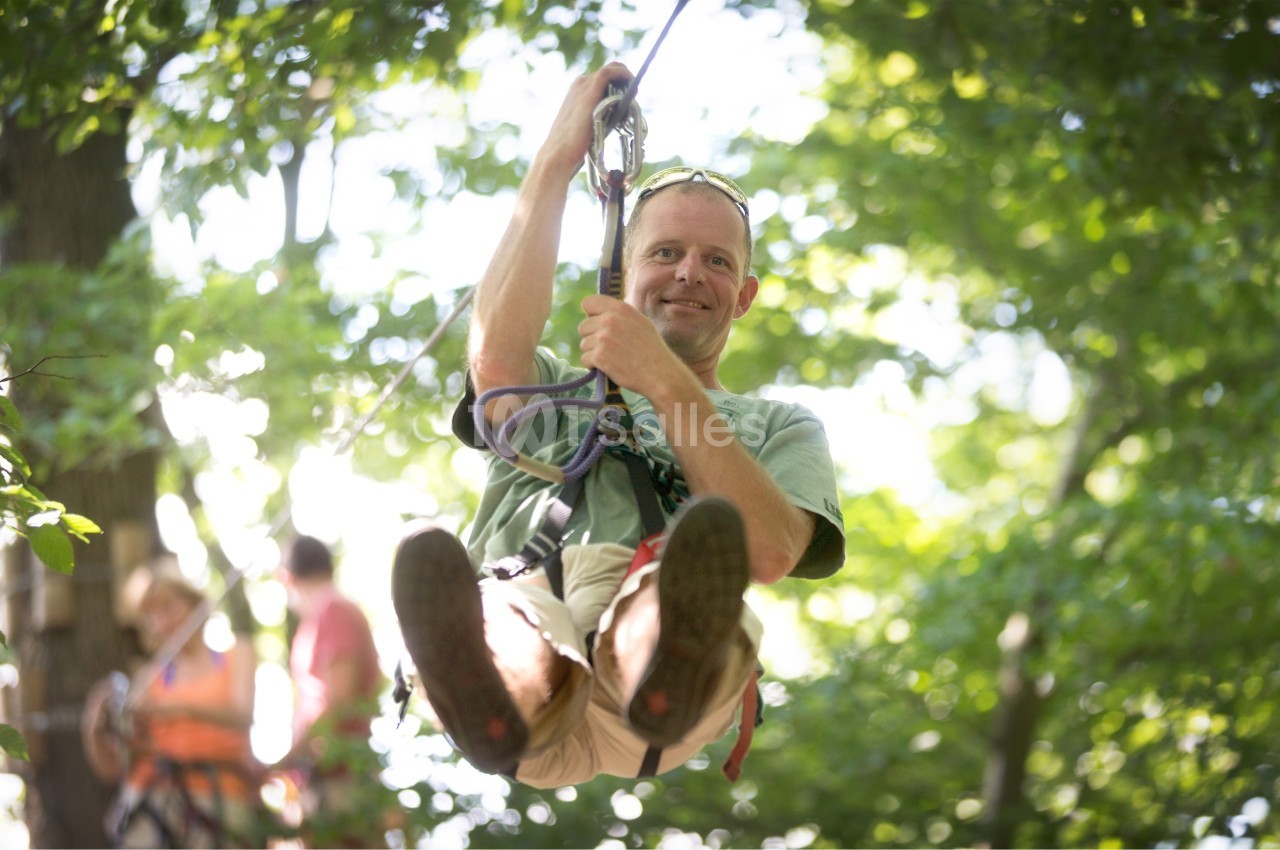 Un homme souriant descend en tyrolienne dans une forêt verdoyante, sous une lumière naturelle.