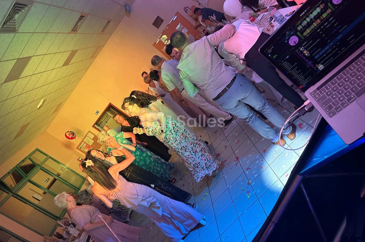 Des personnes dansent et discutent dans une salle éclairée de lumières colorées, avec une table de DJ au premier plan.
