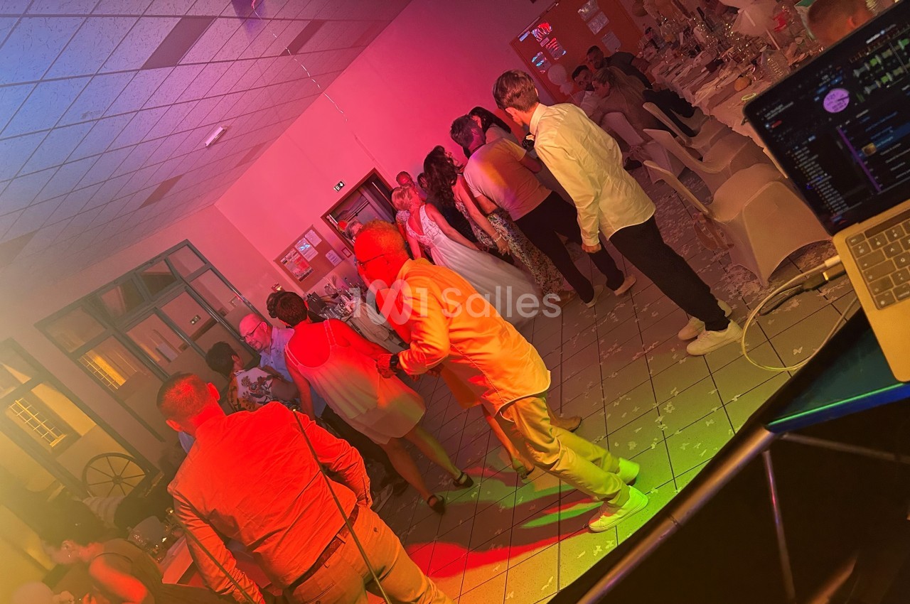 Des personnes dansent et discutent dans une salle éclairée par des lumières colorées, avec une table de DJ au premier plan.