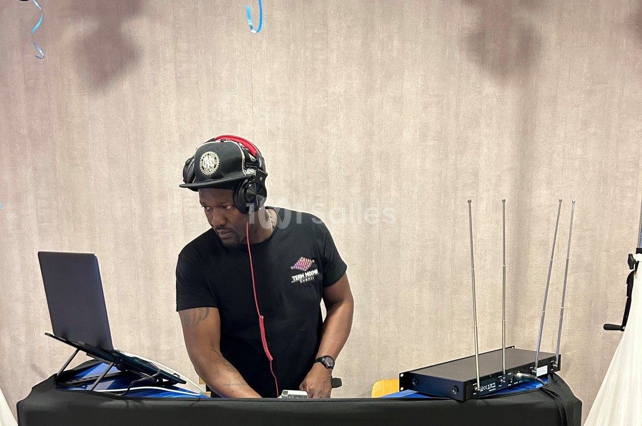 Un DJ mixe de la musique sur un ordinateur portable, portant un casque audio et une casquette, devant un fond neutre.