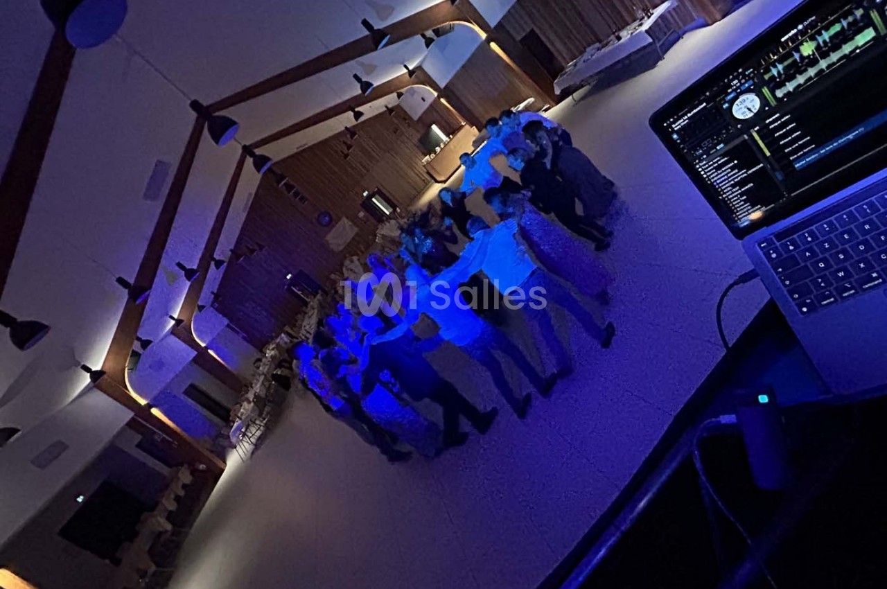 Groupe de personnes dansant en cercle dans une salle éclairée par des lumières bleues, avec un ordinateur de DJ au premier…