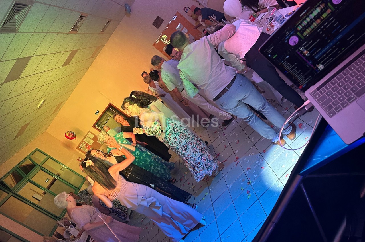 Des personnes dansent et discutent dans une salle éclairée de lumières colorées, avec une table de DJ au premier plan.